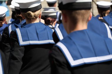 Arkadan yakından bakınca, koyu mavi üniformalı, düz şapkalı, lacivert üniformalı birçok kişi görülüyor. Görünür yüzleri veya özel işaretleri olmayan başları ve omuzlarına verilen ilgi, birlik ve disiplini vurguluyor..