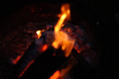 Bir kamp ateşinin samimi ve sıcak parıltısını yansıtan yakın çekim, düşük ışık fotoğrafı. Görüntü yanan odun yığınına odaklanıyor, parlak, dans eden sarı ve turuncu alevler dokulu kabuğu yalıyor.. 
