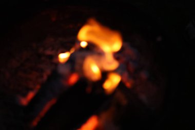 Bir kamp ateşinin samimi ve sıcak parıltısını yansıtan yakın çekim, düşük ışık fotoğrafı. Görüntü yanan odun yığınına odaklanıyor, parlak, dans eden sarı ve turuncu alevler dokulu kabuğu yalıyor.. 