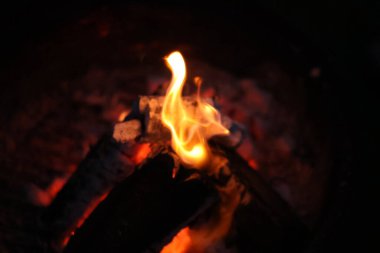 Bir kamp ateşinin samimi ve sıcak parıltısını yansıtan yakın çekim, düşük ışık fotoğrafı. Görüntü yanan odun yığınına odaklanıyor, parlak, dans eden sarı ve turuncu alevler dokulu kabuğu yalıyor.. 
