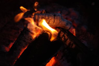 Bir kamp ateşinin samimi ve sıcak parıltısını yansıtan yakın çekim, düşük ışık fotoğrafı. Görüntü yanan odun yığınına odaklanıyor, parlak, dans eden sarı ve turuncu alevler dokulu kabuğu yalıyor.. 