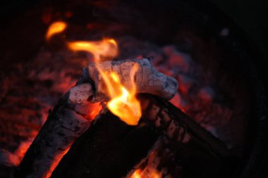 Bir kamp ateşinin samimi ve sıcak parıltısını yansıtan yakın çekim, düşük ışık fotoğrafı. Görüntü yanan odun yığınına odaklanıyor, parlak, dans eden sarı ve turuncu alevler dokulu kabuğu yalıyor.. 