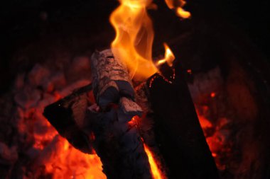 Bir kamp ateşinin samimi ve sıcak parıltısını yansıtan yakın çekim, düşük ışık fotoğrafı. Görüntü yanan odun yığınına odaklanıyor, parlak, dans eden sarı ve turuncu alevler dokulu kabuğu yalıyor.. 