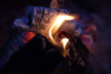 Yakın plan, kömürleşmiş ahşap kütüklerin, parlak sarı alevlerin ve közlerden yayılan pembe ve mor ışığın eşsiz bir fotoğrafı. Görüntü, farklı bir renk paletiyle rüya gibi ve karamsar bir atmosfer yakalıyor.