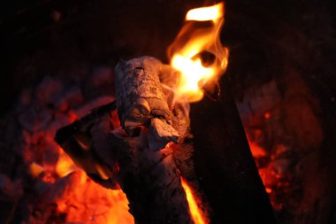 Bir kamp ateşinin samimi ve sıcak parıltısını yansıtan yakın çekim, düşük ışık fotoğrafı. Görüntü yanan odun yığınına odaklanıyor, parlak, dans eden sarı ve turuncu alevler dokulu kabuğu yalıyor.. 