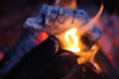 Yakın plan, kömürleşmiş ahşap kütüklerin, parlak sarı alevlerin ve közlerden yayılan pembe ve mor ışığın eşsiz bir fotoğrafı. Görüntü, farklı bir renk paletiyle rüya gibi ve karamsar bir atmosfer yakalıyor.