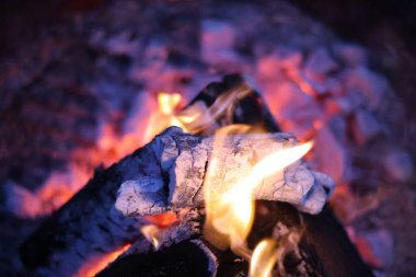 Yakın plan, kömürleşmiş ahşap kütüklerin, parlak sarı alevlerin ve közlerden yayılan pembe ve mor ışığın eşsiz bir fotoğrafı. Görüntü, farklı bir renk paletiyle rüya gibi ve karamsar bir atmosfer yakalıyor.