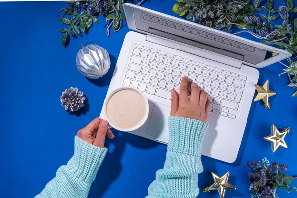 Christmas sale, preparation for holidays concept. Winter office workplace background. Laptop, hot chocolate latte cup with Christmas toys, winter tree branches, flat lay on bright blue background