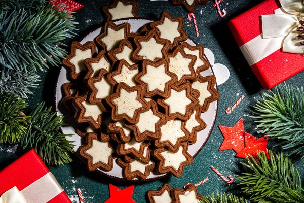 Christmas star sugar glazed cookies, traditional Xmas dark and white chocolate Gingerbread biscuits with holiday decoration, gifts, Christmas tree branch on dark green table top view copy space