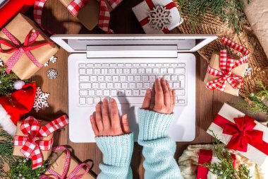 Tahta arka planda Noel hediyesi kutuları olan dizüstü bilgisayar kullanan biri. Kadın eli hediyeli defter, köknar ağacı ve kış dekoru. Noel ve Yeni Yıl için Hazırlık