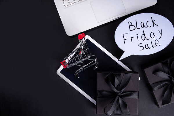 Черная пятница фон распродажи. Simple black flat lay with sign "Black friday", gift boxes, shopping cart, laptop, tablet, headphones top view copy space. Концепция покупок в Черную пятницу