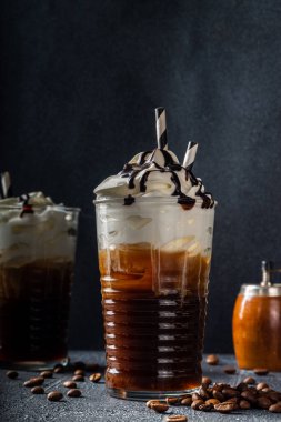 Soğuk kahve içen frappe (frappuccino), krema ve çikolata şurubuyla, pipetlerle, kahve çekirdekleri koyu gri bir taş masada, boşluğu kopyala 