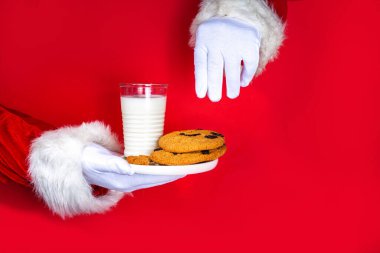 Noel Baba 'nın eli, geleneksel Noel kurabiyesi atıştırmalığıyla süt bardağıyla. Noel Baba Batıran Kurabiye, parlak kırmızı yırtık kağıt delikli arka planda. Mutlu Noeller ve mutlu yıllar tebrik kartı.