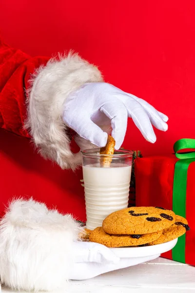 Noel Baba 'nın eli, geleneksel Noel kurabiyesi atıştırmalığıyla süt bardağıyla. Santa Dunking Cookie, parlak kırmızı ve beyaz ahşap arka planda. Mutlu Noeller ve mutlu yıllar tebrik kartı.