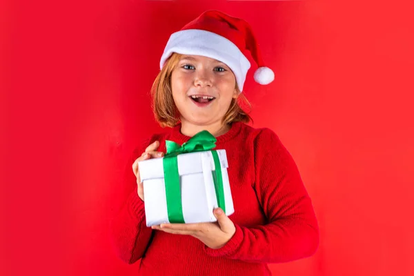 Christmas holiday background with funny blond girl. Little kid girl in red Christmas sweater and funny santa hat costume, with gift box on red background, Xmas greeting card, copy space