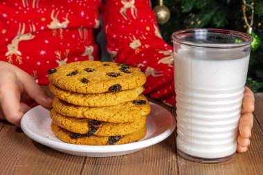 Pijamalı sevimli kız Noel Baba 'ya kurabiye ve süt hazırlıyor. Süslü Noel ağacı arka planına yakın plan. Noel ve hediye konseptini bekliyorum.