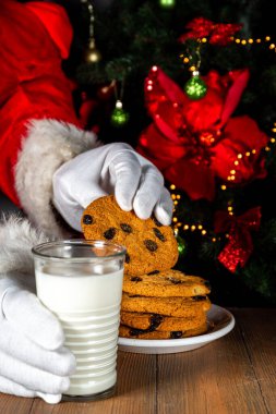 Noel Baba kurabiye ve süt yiyor. Noel Baba elleri Noel ağacına karşı ahşap masada kurabiye ve süt bardağı topluyor., 