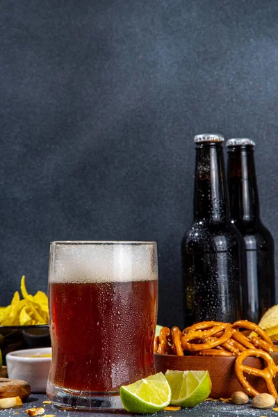 Beer with various salted snacks set. Black table background with traditional party snacks, beer bottles and glasses, with chips, onion rings, salted nuts, crisps and sauces top view copy space