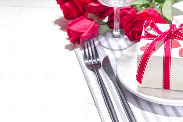 Valentine day table setting with plate, gift box with festive red ribbon, wine glass, fork and knife, red roses flowers bouquet white tiled table flatlay copy space