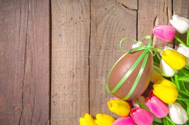 Paskalya tebrik kartı arka planında kocaman bir çikolata yumurtası, şenlikli lale çiçekleri buketi, güneşle aydınlatılmış ahşap arka plan fotokopi alanı.