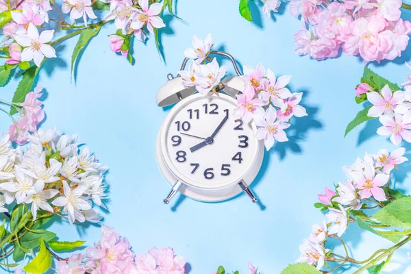 Spring Time Change background. Beautiful fresh spring tree flowers with alarm clock on light blue sunny background top view copy space