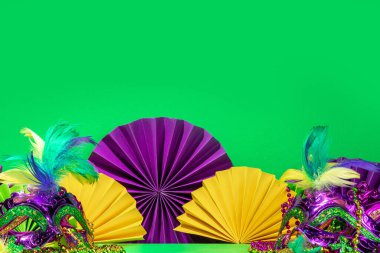 Mardi Gras colourful holiday greeting card background with festival masquerade accessories, decor, carnival mask, beads, feathers, fan on bright background traditional yellow purple green colors