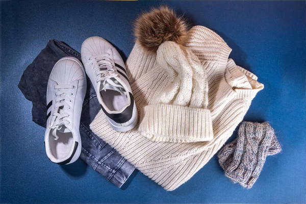 Set of female winter autumn clothes. Jeans, white knitted sweater and hat,  mittens and sneakers on  dark blue background. Casual cozy fashion outfit flat lay