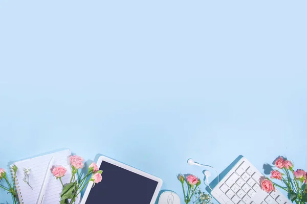 Spring office desk flatlay with keyboard, notepad, tablet, mouse, headphones, spring flowers and leaves, high-colored bright sunny blue background, top view frame