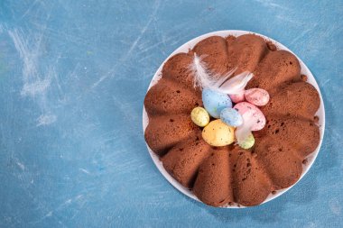 Traditional cake for Easter. Chocolate and vanilla Easter round cake with chocolate and sugar powder sprinkles, colorful Easter eggs, spring flowers, on a blue background copy space
