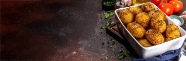 Homemade hasselback potatoes it baking tray. Baked hasselback potatoes with cheese, garlic and herbs, on kitchen table with ingredient for cooking