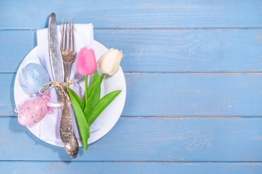 Easter table setting with tulip flowers and eggs, tag with text Happy Easter, over light blue wooden background top view copy space