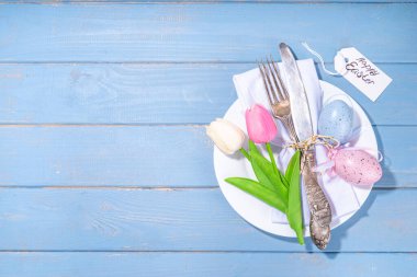 Easter table setting with tulip flowers and eggs, tag with text Happy Easter, over light blue wooden background top view copy space