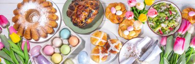Festivalde Paskalya yemeği veya brunch masası sunuldu, Paskalya yumurtası, lale çiçeği buketi, geleneksel Paskalya yemekleri - pasta, çapraz çörekler, çelenk ekmeği, jöleli jambon ve bahar salatası, beyaz masa arkaplanı