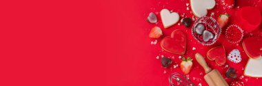 High-colored bright red Valentine day baking background, Various bakery ingredients, cookies, chocolates, sugar sprinkles, strawberry, rolling pin and whisk, for cooking valentine`s sweets, desserts