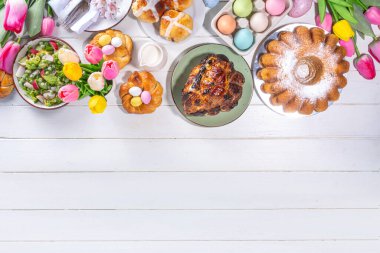 Festivalde Paskalya yemeği veya brunch masası sunuldu, Paskalya yumurtası, lale çiçeği buketi, geleneksel Paskalya yemekleri - pasta, çapraz çörekler, çelenk ekmeği, jöleli jambon ve bahar salatası, beyaz masa arkaplanı