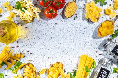 Beyaz masa arkasında çeşitli kurutulmuş spagetti, makarna ve kaşıklarla hazırlanmış makarna. Farklı şekillerde çiğ İtalyan makarnası, otlar, zeytinyağı, domatesler, baharatlar.