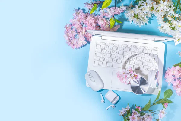 Spring office workplace, blogging flat lay background. White laptop, with headphones, tablet, spring flowers bouquet on light blue background, girl\'s hands typing on a laptop.