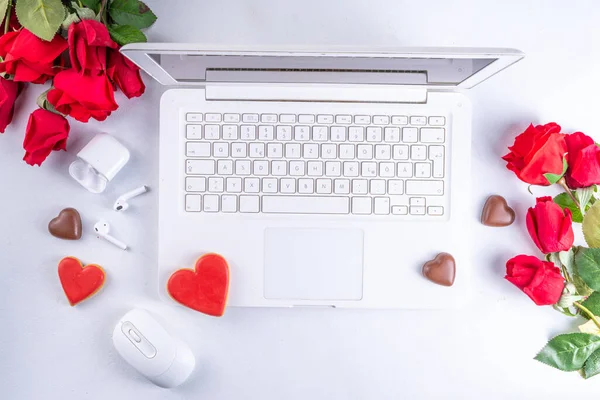 Woman using laptop with red roses. White laptop on white table background with chocolates, cookie hearts, red roses bouquet, flat lay working holiday, Valentine day background, top view copy space