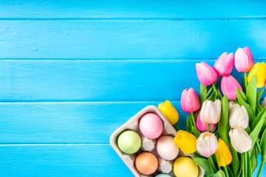 Simple minimal Easter greeting card background, colorful pastel Easter eggs with tulip flower bouquet, on light blue wooden plank background top view copy space