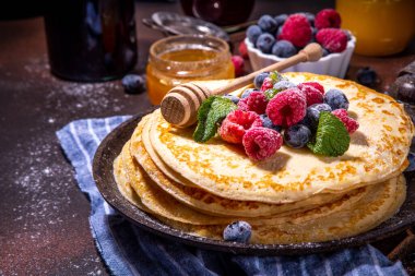 Homemade french tiny pancakes, battercakes with berry. Stack of freshly baked thin crepes on frying pan with fresh blueberry, raspberries and honey. Healthy morning breakfast on dark background