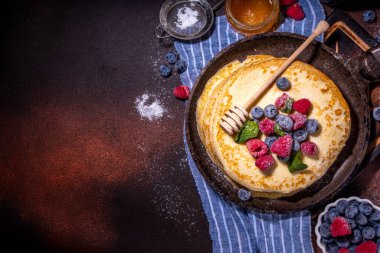 Homemade french tiny pancakes, battercakes with berry. Stack of freshly baked thin crepes on frying pan with fresh blueberry, raspberries and honey. Healthy morning breakfast on dark background