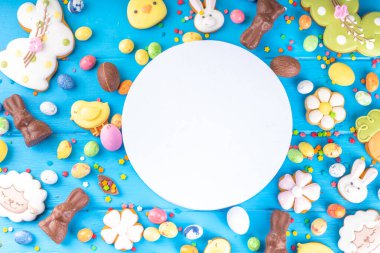 Easter sweets and candies background. High-colored blue flat lay with various Easter chocolate eggs, treats, symbols of easter sheep, bunny, carrot, flowers, cookies, top view copy space, frame
