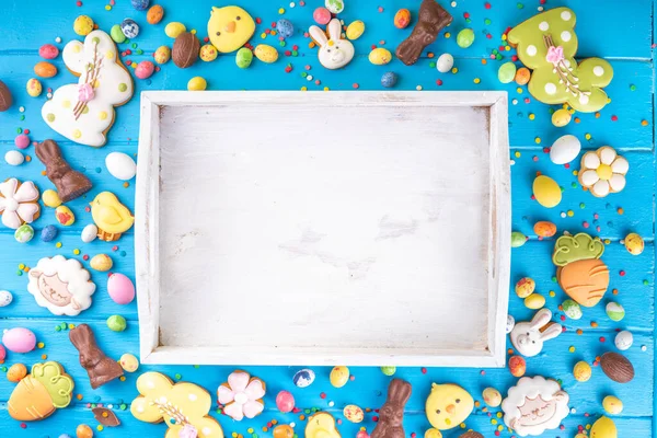 Easter sweets and candies background. High-colored blue flat lay with various Easter chocolate eggs, treats, symbols of easter sheep, bunny, carrot, flowers, cookies, top view copy space, frame