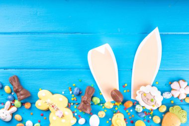 Easter sweets and candies background. High-colored blue flat lay with various Easter chocolate eggs, treats, symbols of easter sheep, bunny, carrot, flowers, cookies, top view copy space, frame