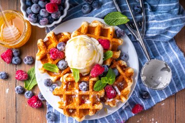 Tasty Homemade Belgian waffles with raspberries, blueberries, vanilla ice cream and syrup or honey, sweet summer healthy breakfast, light wooden background copy space 