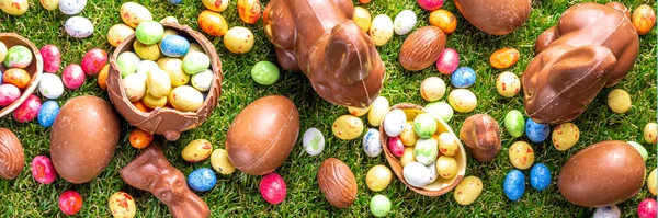 Easter egg hunting background. Various candy and chocolate Easter eggs, bunny and rabbits with basket for eggs on green grass park or garden background