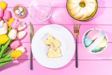 Şirin Paskalya sofrası, bahar tatili tabağı, cam, çatal bıçak takımı, lale çiçeği buketi, Paskalya dekorasyon sembolleri ve ikramlar, pembe ahşap parlak masa, üst görünüm kopya alanı.