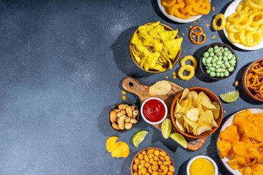 Various unhealthy salty bar snacks set. Dark grey table background with traditional party snacks - chips, onion rings, salted nuts, crisps,  pretzels and sauces top view copy space