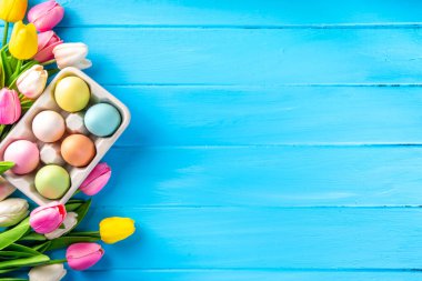 Simple minimal Easter greeting card background, colorful pastel Easter eggs with tulip flower bouquet, on light blue wooden plank background top view copy space