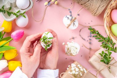 Eco friendly zero waste Easter flatlay. Spring flowers, leaves, eggs on pink pastel background, with craft paper gift box. Woman hands decorate eggs with organic natural decor, preparation for Easter